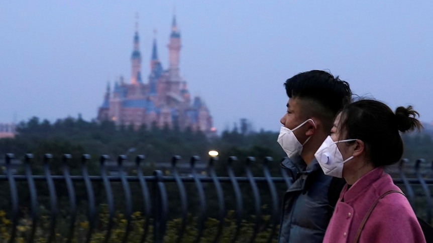 Shangai Disney castle turrets in background with a couple in facemasks out-of-focus in foreground