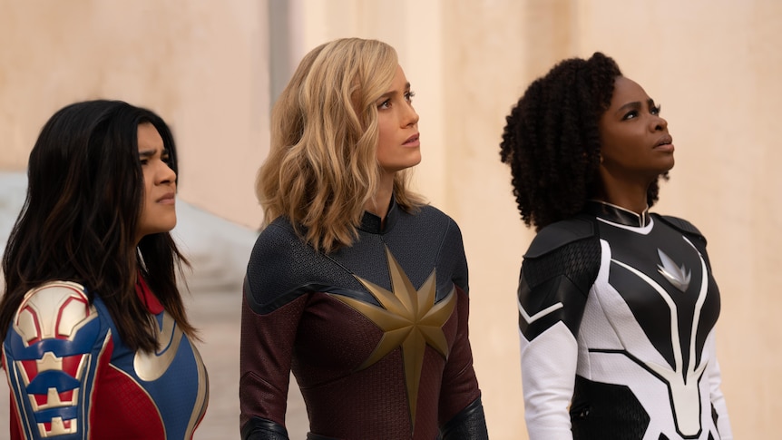 A brunette Pakistani woman, a blonde white woman and a dark brunette black woman wearing superhero outfits stand looking skyward