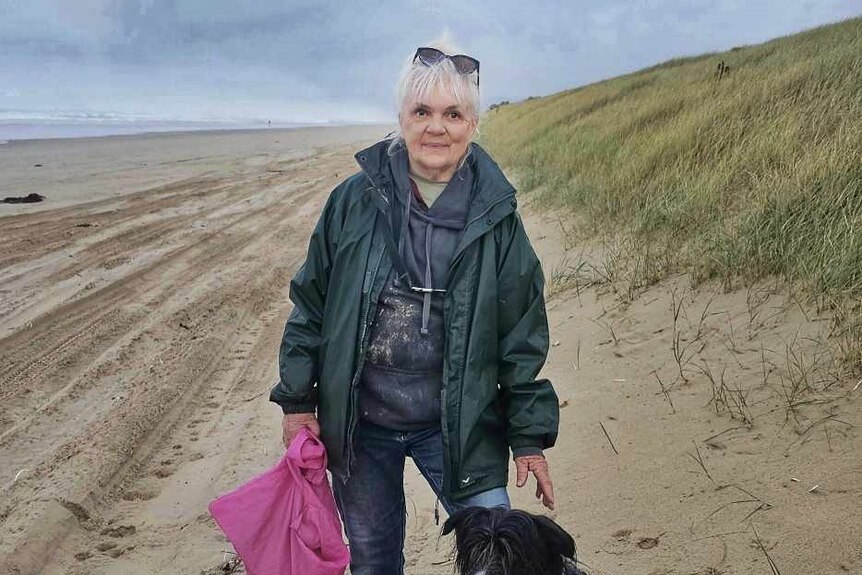 A woman holding a pink bag stands on a beach next to a wet and sandy black and white dog