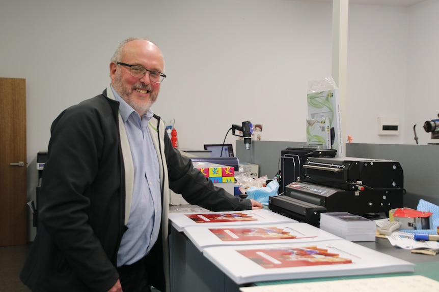 Mr Gates stands in a busy printing workshop, over looking pile of big red posters.