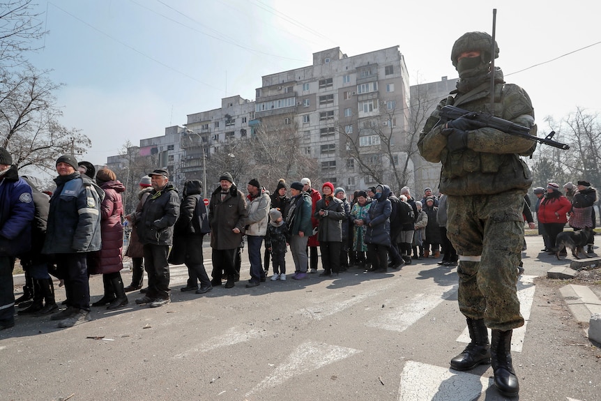 A Russian army soldier stands next to local residents in line for aid.