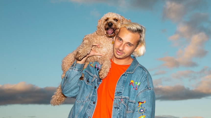 Perth producer Flume poses in an orange shirt and blue denim jacket. His golden retriever cross poodle is on his shoulder.