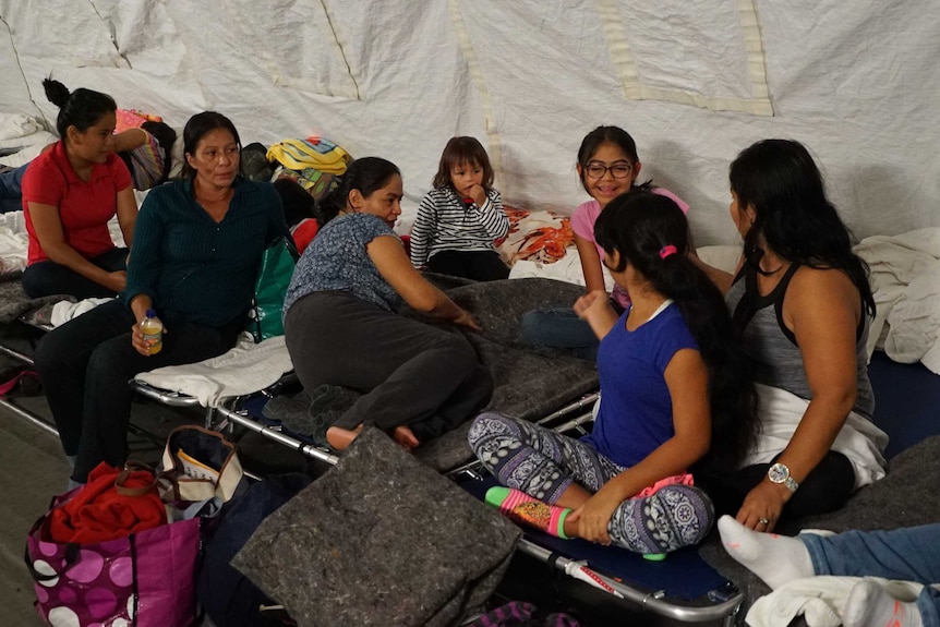 A group of migrants from Mexico — made up of women and children — sit on camp beds in a tent.