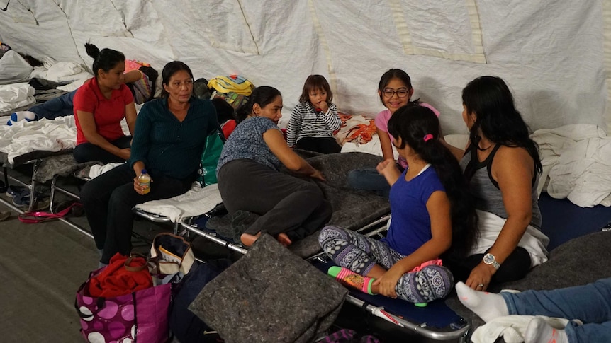 A group of migrants from Mexico — made up of women and children — sit on camp beds in a tent.