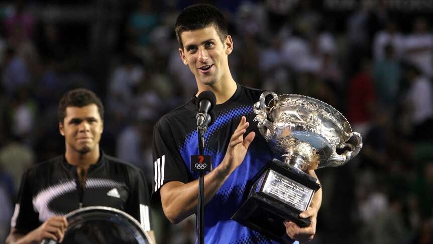 Novak Djokovic (front) holds the Australian Open trophy while speaking into a microphone. Jo-Wilfried Tsonga watches on.