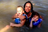 Tanya Day, smiling, holding two children wearing blue flotation vests, in the brown water of the Murray river.