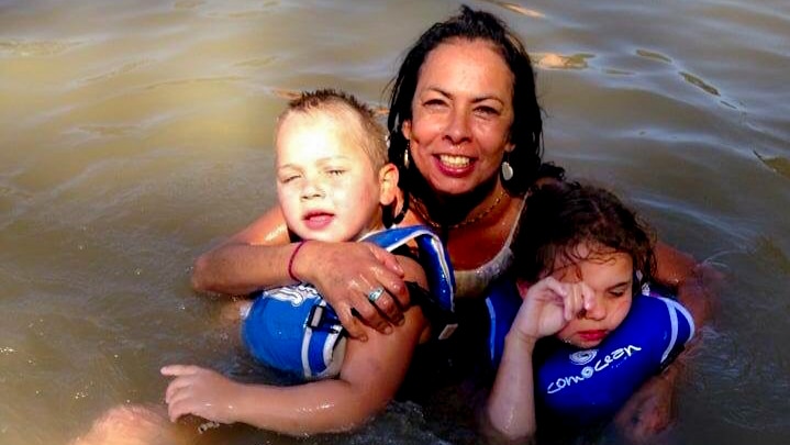 Tanya Day, smiling, holding two children wearing blue flotation vests, in the brown water of the Murray river.