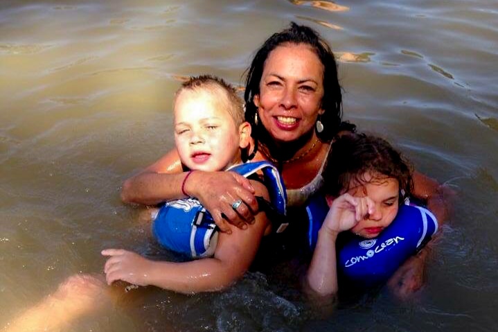 Tanya Day, smiling, holding two children wearing blue flotation vests, in the brown water of the Murray river.