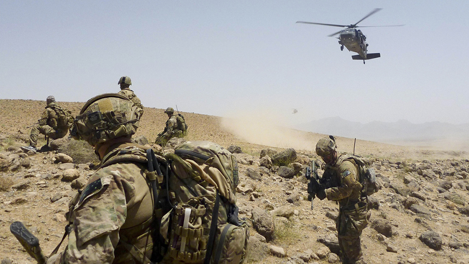 Five soldiers carrying weapons walk across a dusty Afghanistan mound as a helicopter hovers closely above.