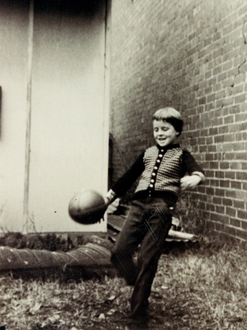 A black and white photo of a child kicking a football in front of a brick wall.