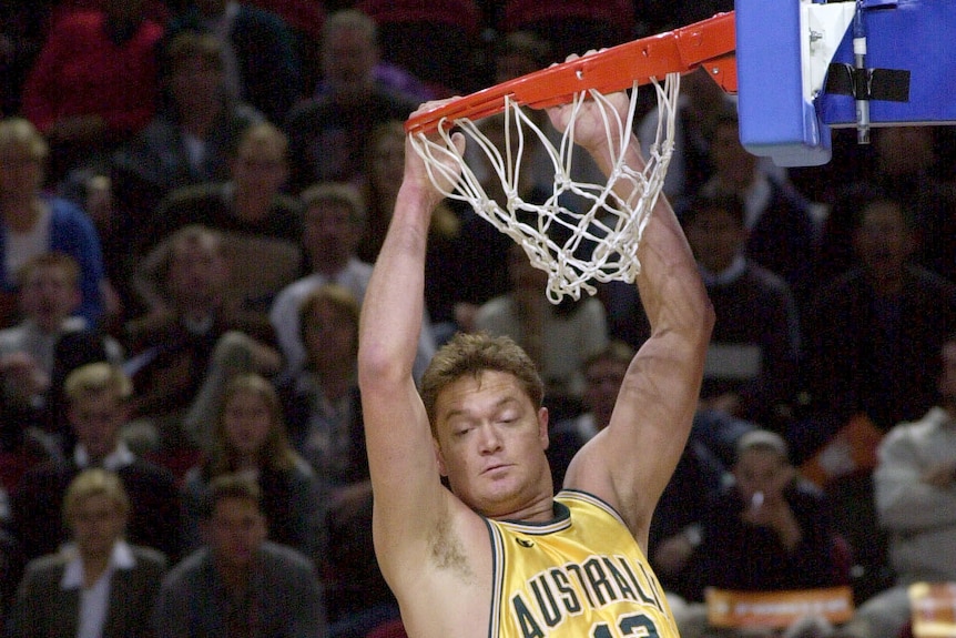 An Australian basketballer slam dunks, two Canadian players look up and watch on underneath