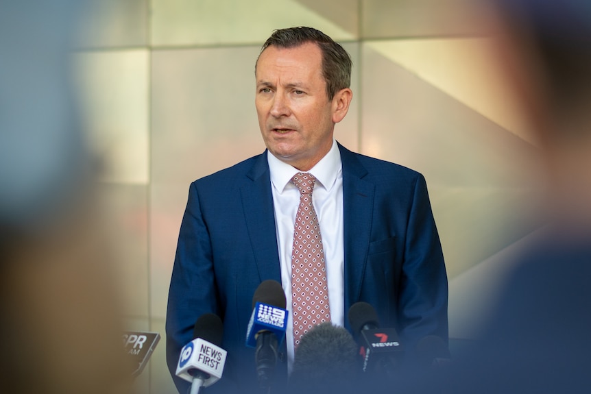 A middle aged man wearing blue suit, white shirt and patterned tie standing in front of light yellow wall.