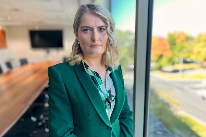 A serious-looking woman in a green suit jacket and patterned green shirt in her stands beside an boardroom window.