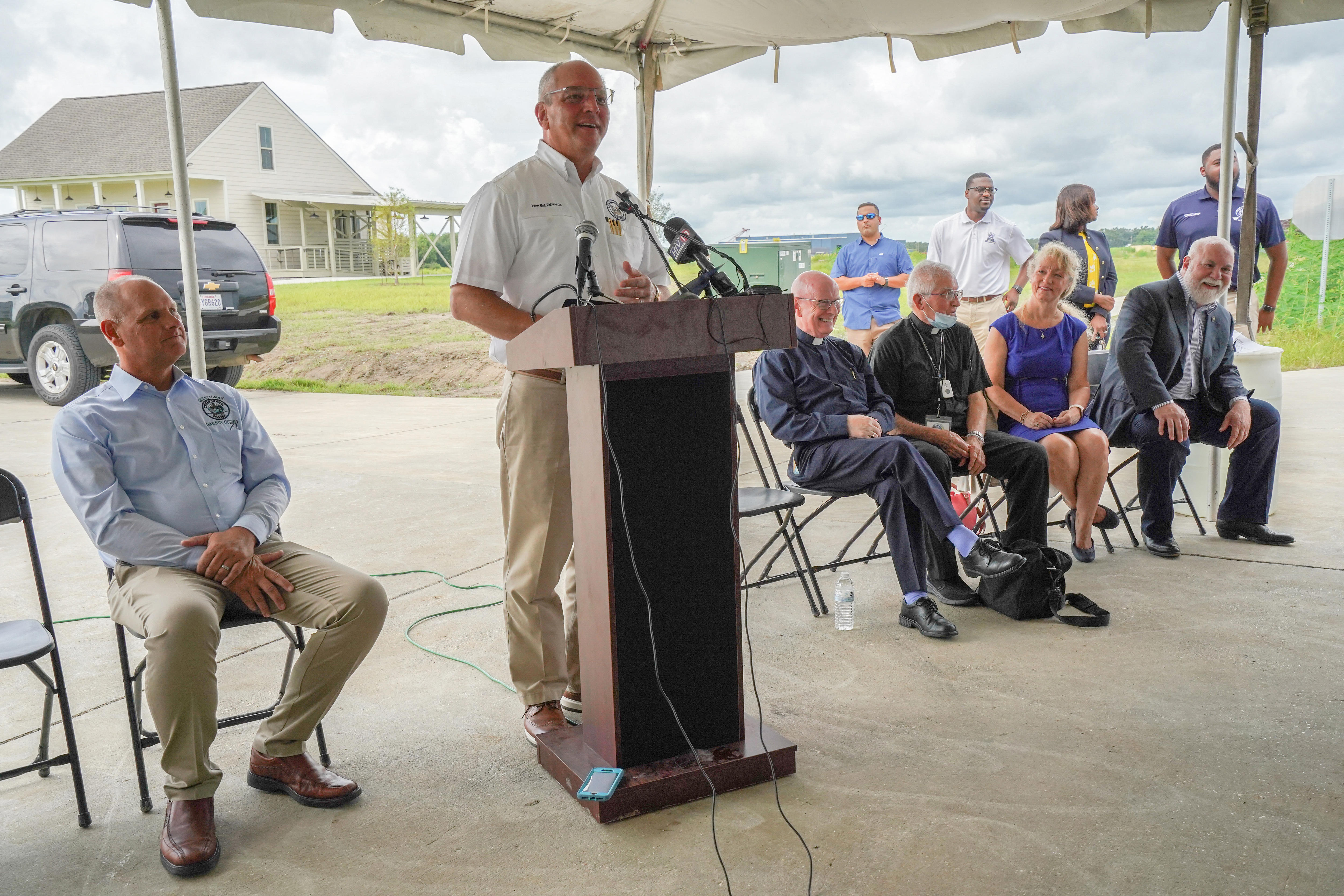 Louisiana Governor John Bel Edwards speaks at a lectern outdoors under a covering as people sit on either side of him