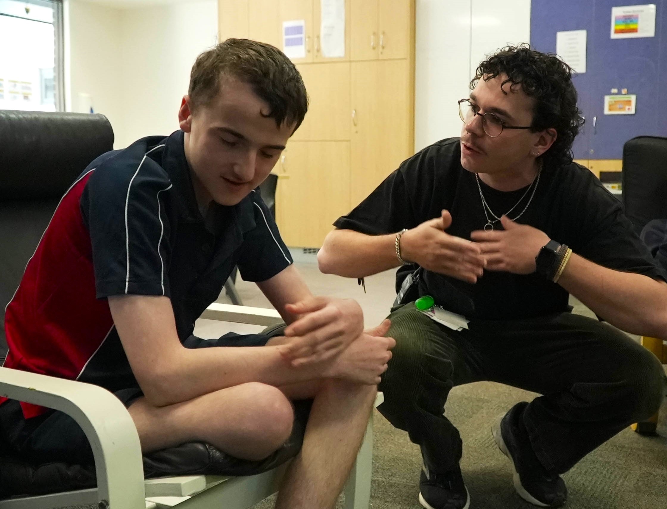 A young white boy with intellectual disability. He is in a classroom and being spoken to by a teacher's aide