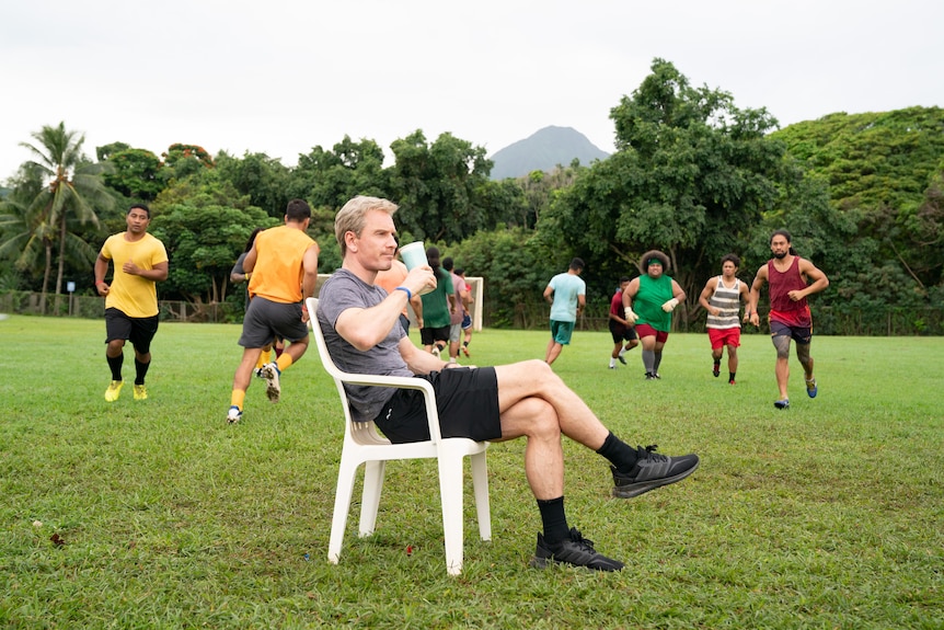 A film still of Michael Fassbender perched on a white plastic chair, as Pacific Islander soccer players practice behind him