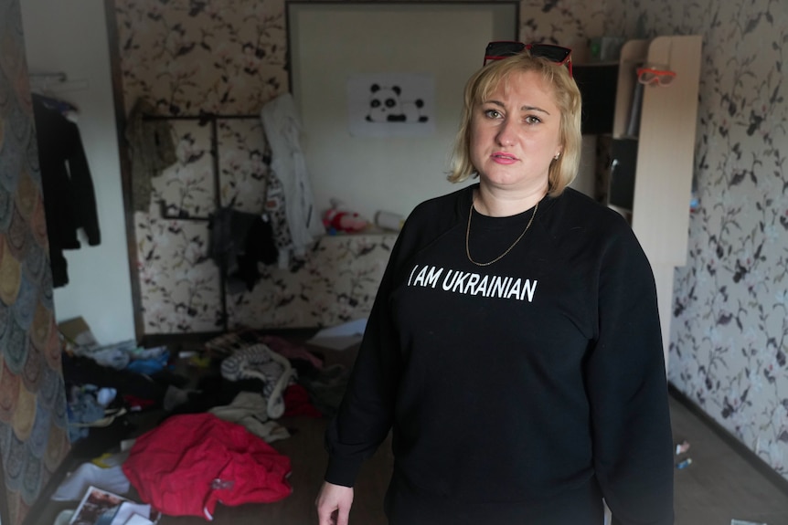 A blonde woman in a black top with "I am Ukrainian" written on it stands in a trashed room