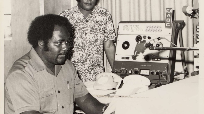 One person sitting at a table with sound recording equipment holding some paper, another stands beside them.