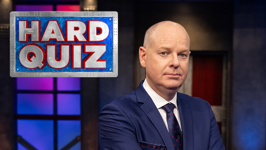 Tom Gleeson standing with his arms crossed with a serious look on his face with th Hard quiz logo in the top left corner