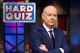 Tom Gleeson standing with his arms crossed with a serious look on his face with th Hard quiz logo in the top left corner