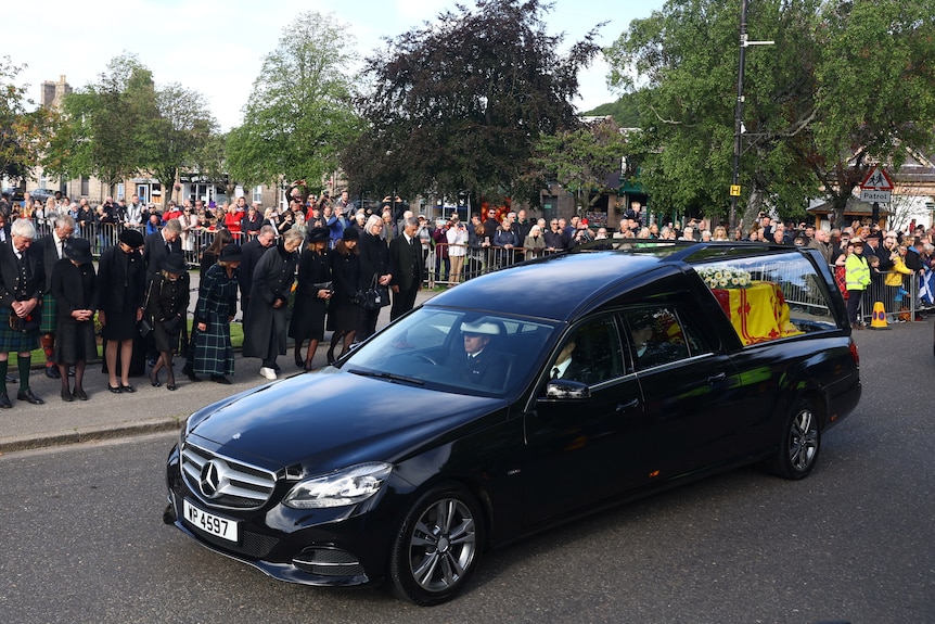 Crowds of people dressed in black bow their heads as the hearse drives by 