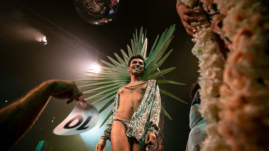 A man in leopard print shorts and long-sleeved shirt smiles on stage at Sissy Ball 2019, under a disco ball.