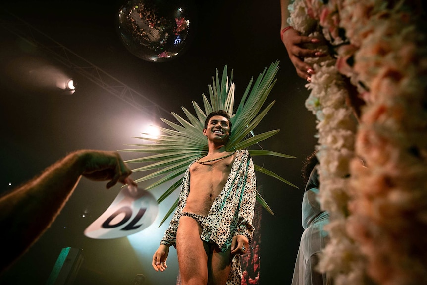 A man in leopard print shorts and long-sleeved shirt smiles on stage at Sissy Ball 2019, under a disco ball.