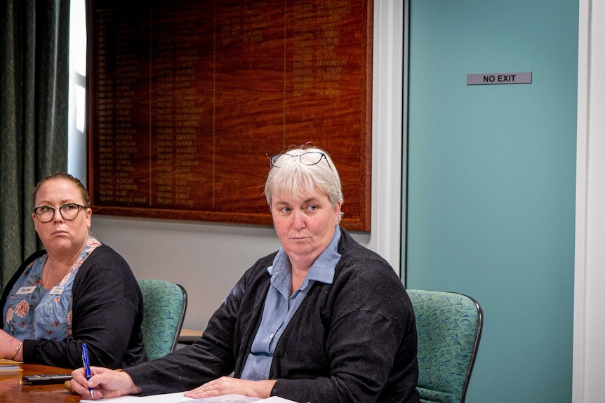 Two women sit at a meeting table with a door saying 'no exit' behind them