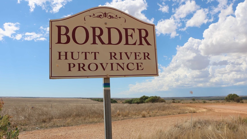 A white and red sign reading 'Border Hutt River Province' stands next to a gravel road under a blue cloudy sky.