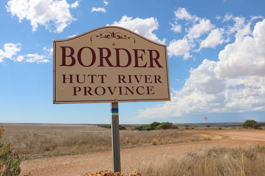 A white and red sign reading 'Border Hutt River Province' stands next to a gravel road under a blue cloudy sky.