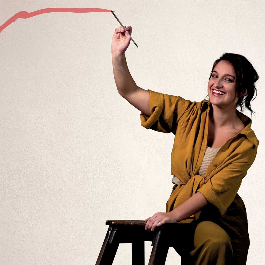 A smiling woman stands on a small ladder. She has painted a thinning line across the wall behind her.
