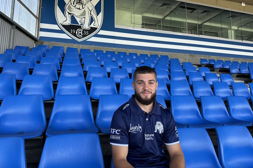 A bearded man wearing a Bulldogs polo shirt sits in blue stadium seats in front of a large Bulldogs logo