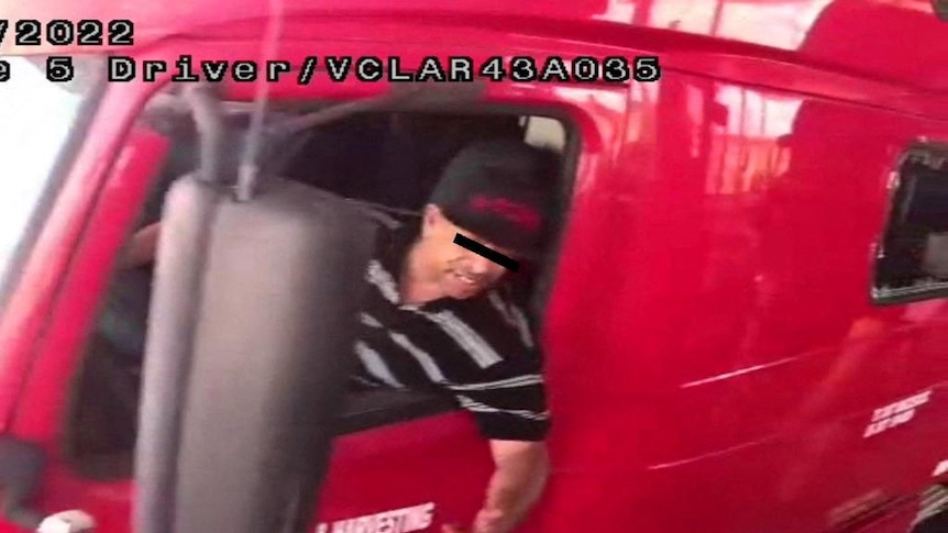 A security camera image of a man wearing a t-shirt and hat leaning out of the window of a large red truck.