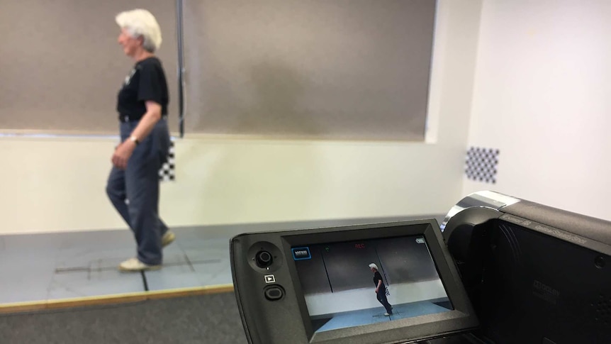 A woman walks on a plastic surface marked with lines and is filmed by small video camera on a tripod.