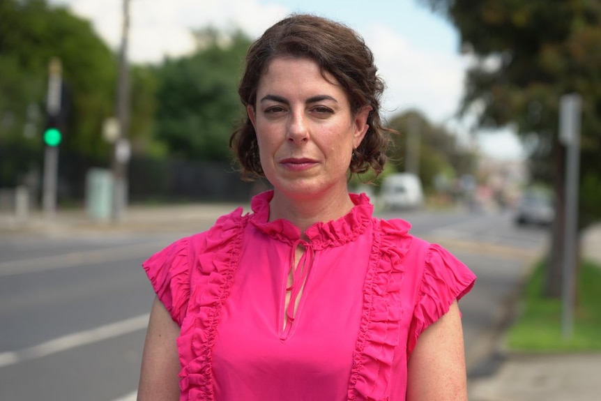 Clare Walter stands in a pink top looking into the camera, while standing next to a busy road