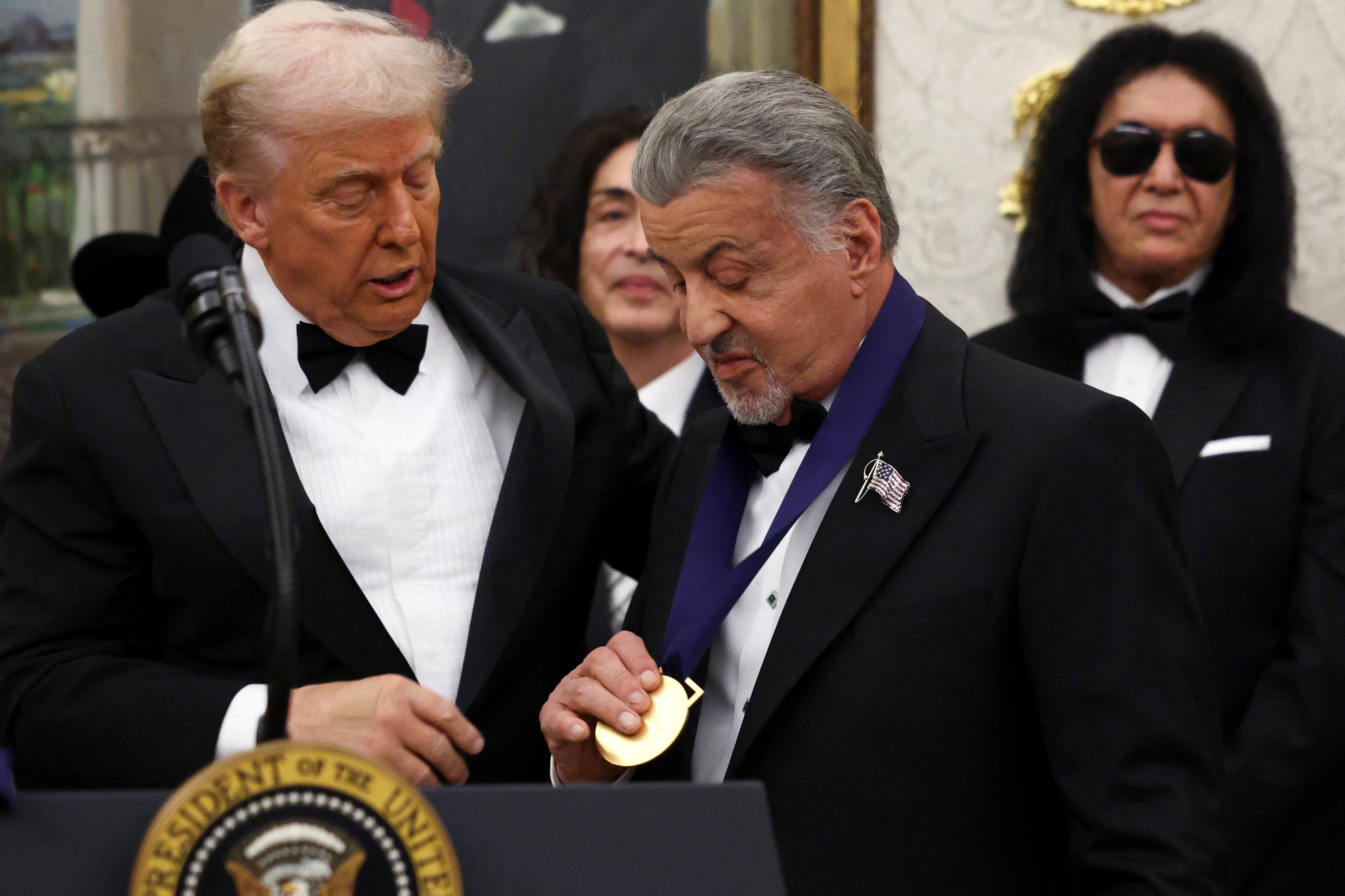 Sylvester Stallone holds a medal while Donald Trump looks on.