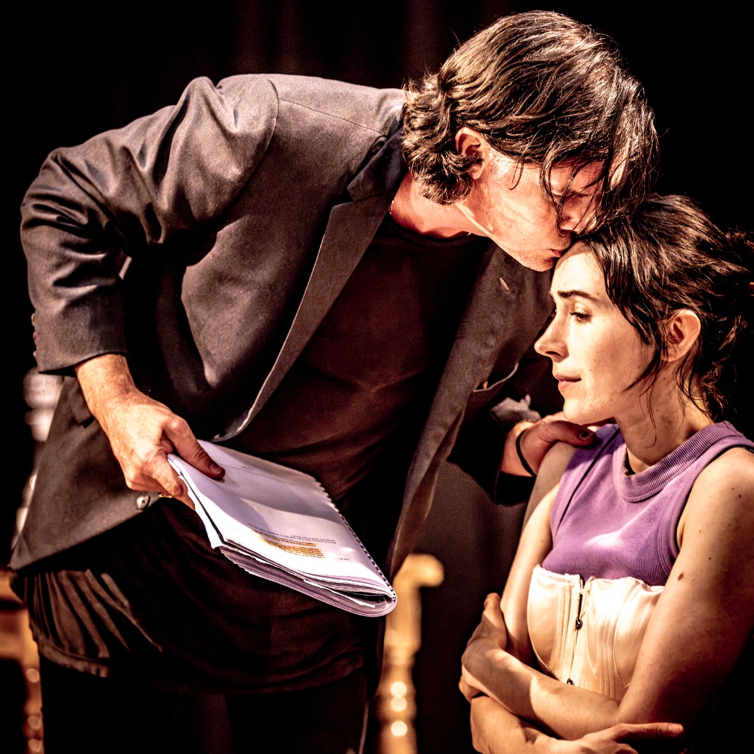 A middle-aged white man kisses a 30-something white woman on the forehead will holding a play script.