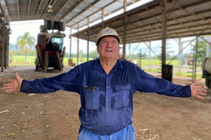 A man in a work shirt and hard hat stands arms open in a shed