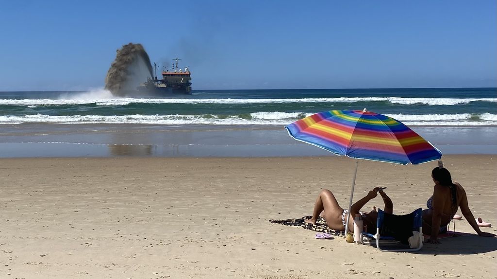 People relaxing on the beach as a barge pumps sand in the distance.