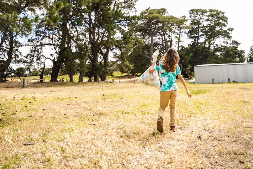 A boy with long hair walks across a grassy field with a bag slung over his shoulder.