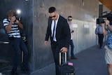 A man in a dark suit and glasses bows his head while being photographed and filmed.