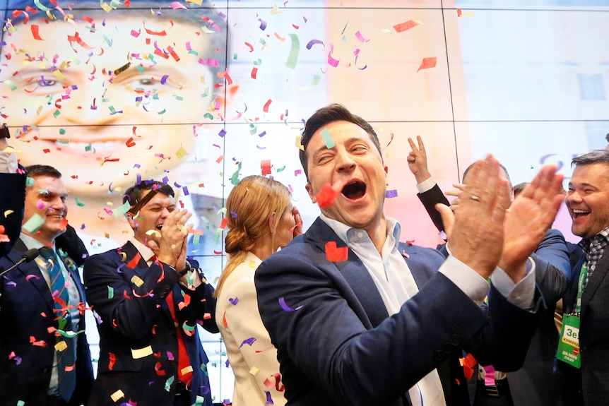 Volodymyr Zelenskyy claps his hands as supporters celebrate behind him and confetti is dispersed into the air