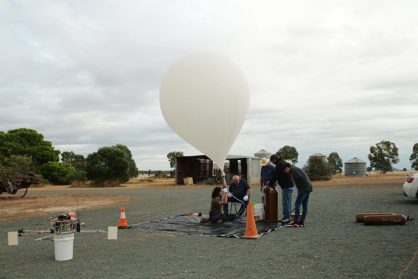 An almost full balloon floats in the air above five people keeping it steady.