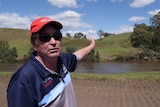 A farmer wearing sunglasses and a red hat gestures at some hills behind him.