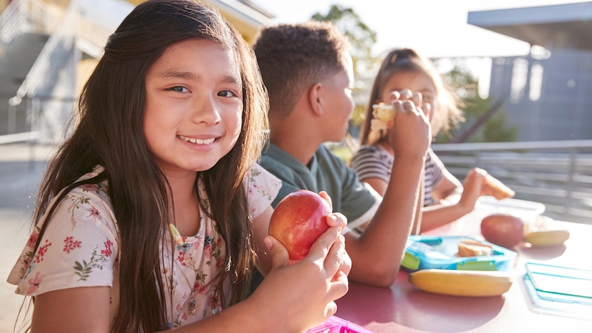 A smiling young female student sits at an outdoor table at school and holds and apple.