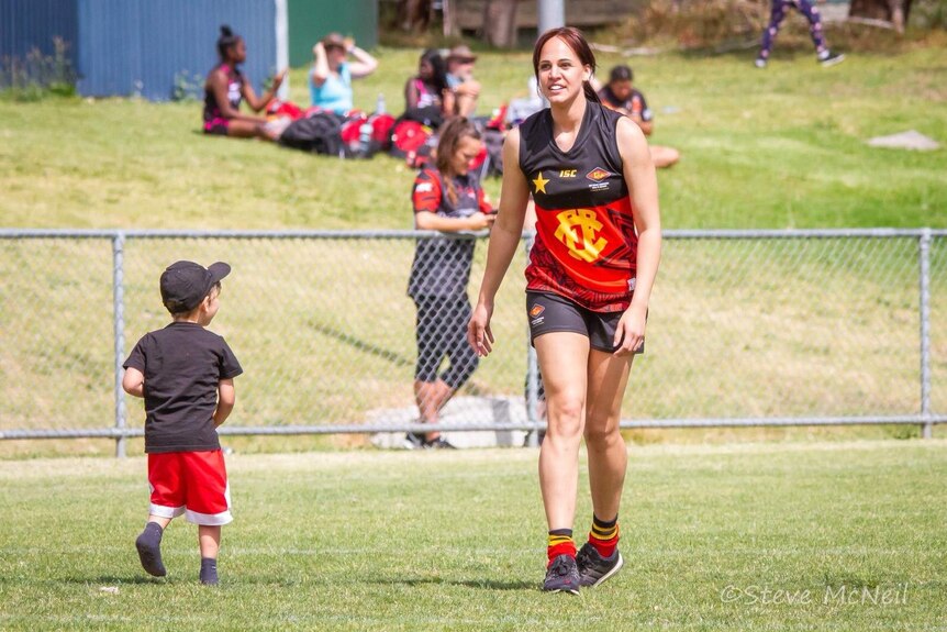 Kirra wearing an AFL uniform walking on a field with her small son Kai