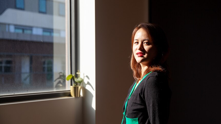 You view a woman of Asian descent half-framed by sunlight while standing next to a bright window.