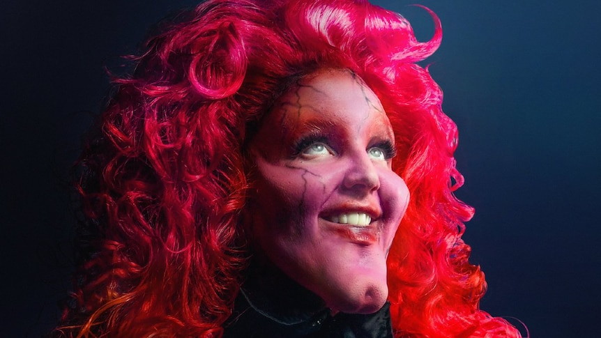Female with chin prosthetics and pink/purple makeship, wearing a long pink neon wig