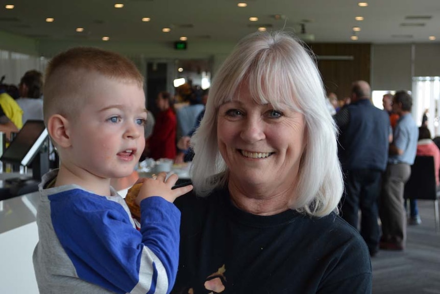 A smiling woman with white hair holds a young boy with blue eyes and blonde hair in a crowded conference room.