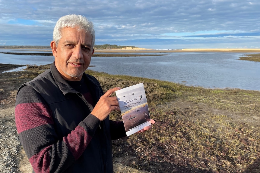 Indigneous elder holds a copy of his book with Lake Tyers in the background